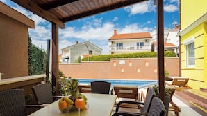 Eine Terrasse im Freien mit einem Esstisch, Korbstühlen und Blick auf einen Swimmingpool und Gebäude im Hintergrund.