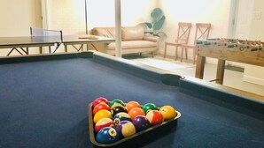A pool table with a set of billiard balls, a foosball table, and a sofa in a room with white walls and a door. - NEW! ARK Family Retreat w/ Game Room 3min to Ark! (Williamstown)