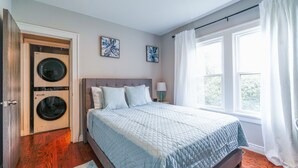 A bedroom with a bed, two wall-mounted washing machines, a window with curtains, and two framed pictures on the wall. - Faboulous and Cozy Apartment in Montclair, NJ (Montclair)