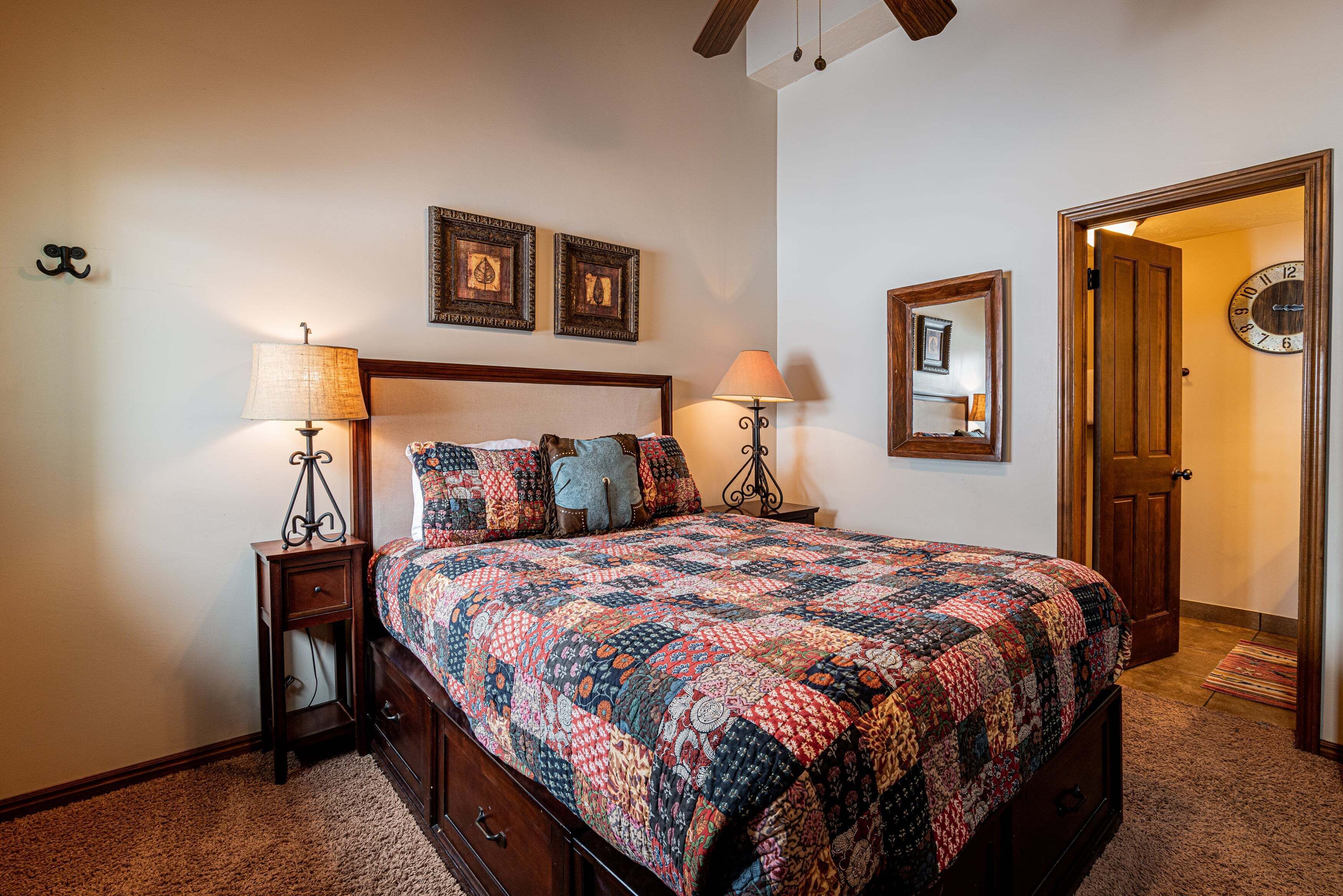 A bedroom with a wooden bed, two bedside tables with lamps, a wall clock, and framed pictures.