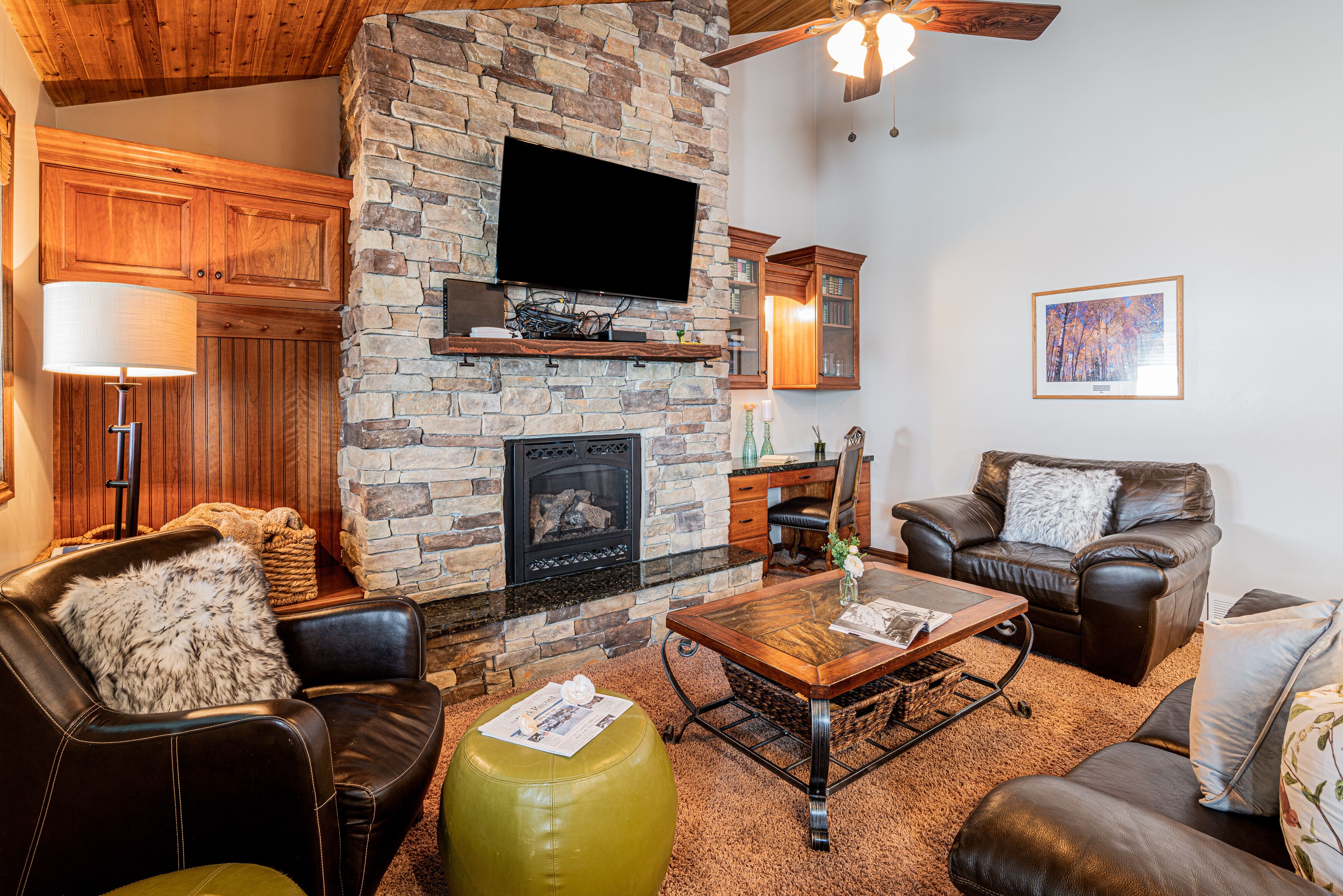 A cozy living room with a stone fireplace, leather furniture, and a flat-screen TV mounted on the wall.