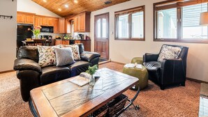 A spacious living room with a wooden coffee table, leather sofa, and armchairs. A kitchen with wooden cabinets and a black refrigerator is visible in the background. - Park City Motherlode F is in a Great Location with Year-Round Recreation! (Park City)
