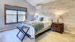 A bedroom with a bed, a window, a wall-mounted air conditioner, a bedside lamp, and a wooden nightstand. - Hot Tub, Decks & More! Serene Maggie Valley Home (Maggie Valley)