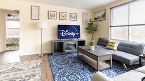 A living room with a flat-screen TV displaying the Disney+ logo, a grey sofa with a 'BOWDY' pillow, a coffee table, a patterned rug, and a potted plant.