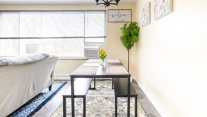 A modern living room with a sofa, a dining table with benches, a chandelier, and a large window with blinds.