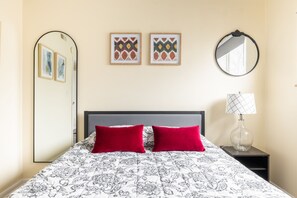 A neatly made bed with a patterned bedspread and two red pillows, a bedside table with a lamp, and two framed artworks on the wall.