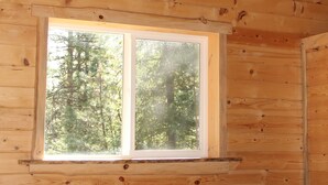 A wooden cabin room with a bed, a window with a view of trees, and a sloped ceiling with a light fixture.