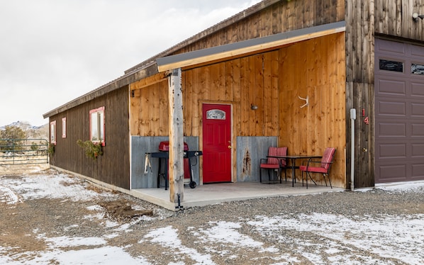 Un bâtiment rustique avec une porte rouge, un espace extérieur abrité avec deux chaises, et un sol de gravier parsemé de plaques de neige.