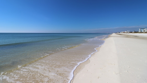 A beach with clear blue water, sandy shore, and buildings in the background.