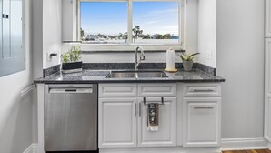 A kitchen with a built-in dishwasher, a double sink, and a window with a view of palm trees and houses.