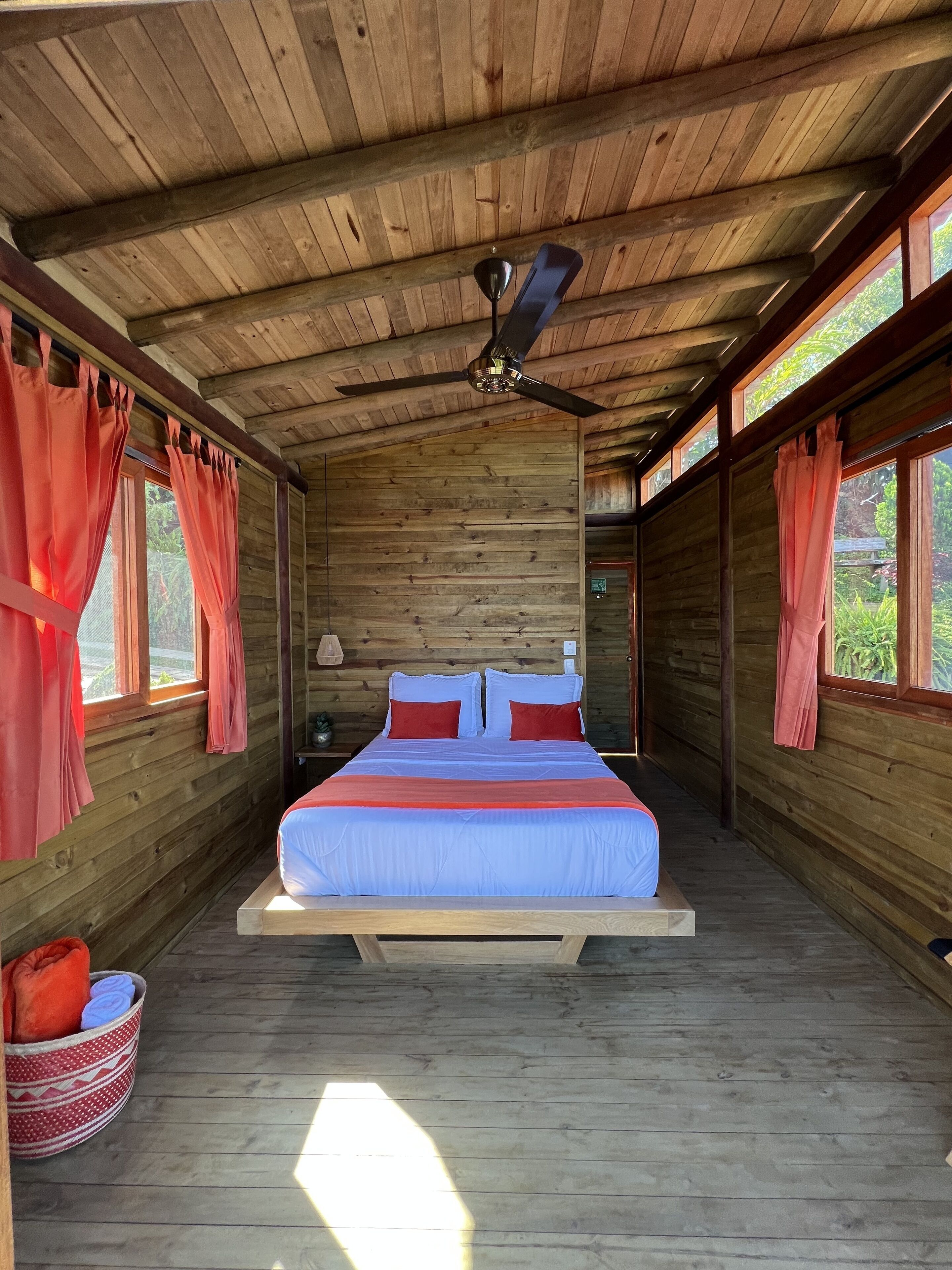 Une chambre dans une cabane en bois, avec un lit, des rideaux et un ventilateur de plafond.