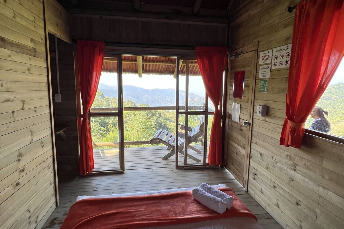 Une chambre rustique en bois, avec un lit, des rideaux rouges et une vue sur les montagnes par une grande fenêtre.