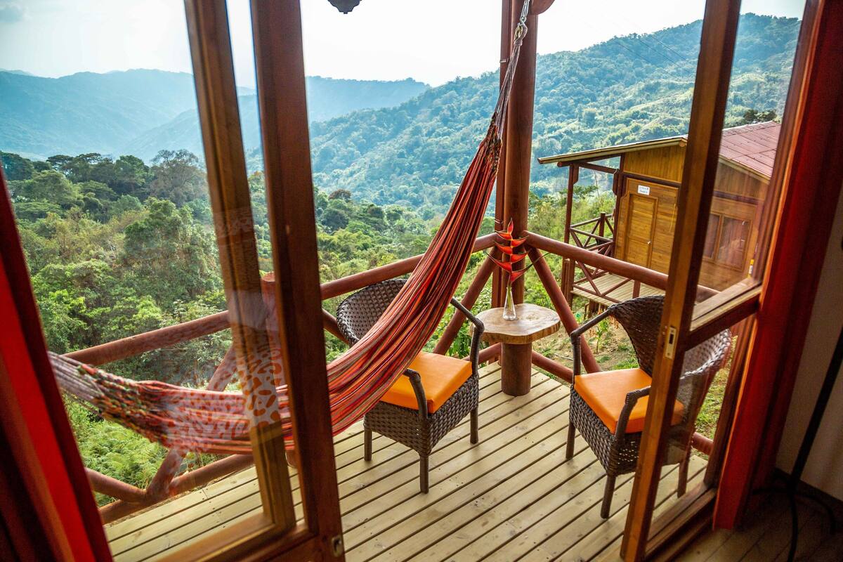 Une terrasse en bois, dotée de chaises en osier et d’un hamac, qui offre une vue imprenable sur une vallée verdoyante.