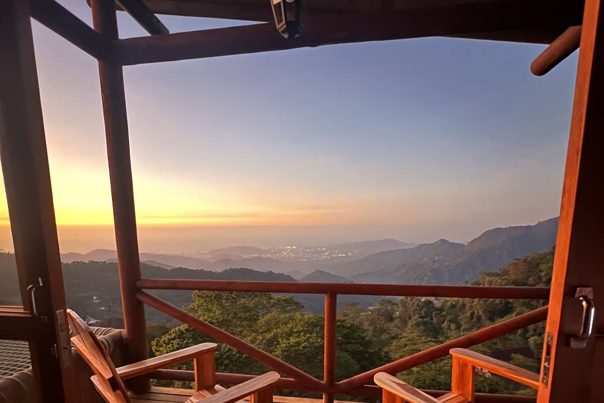 Une terrasse en bois avec deux chaises et une table, d’où l’on peut admirer un paysage montagneux au coucher du soleil.