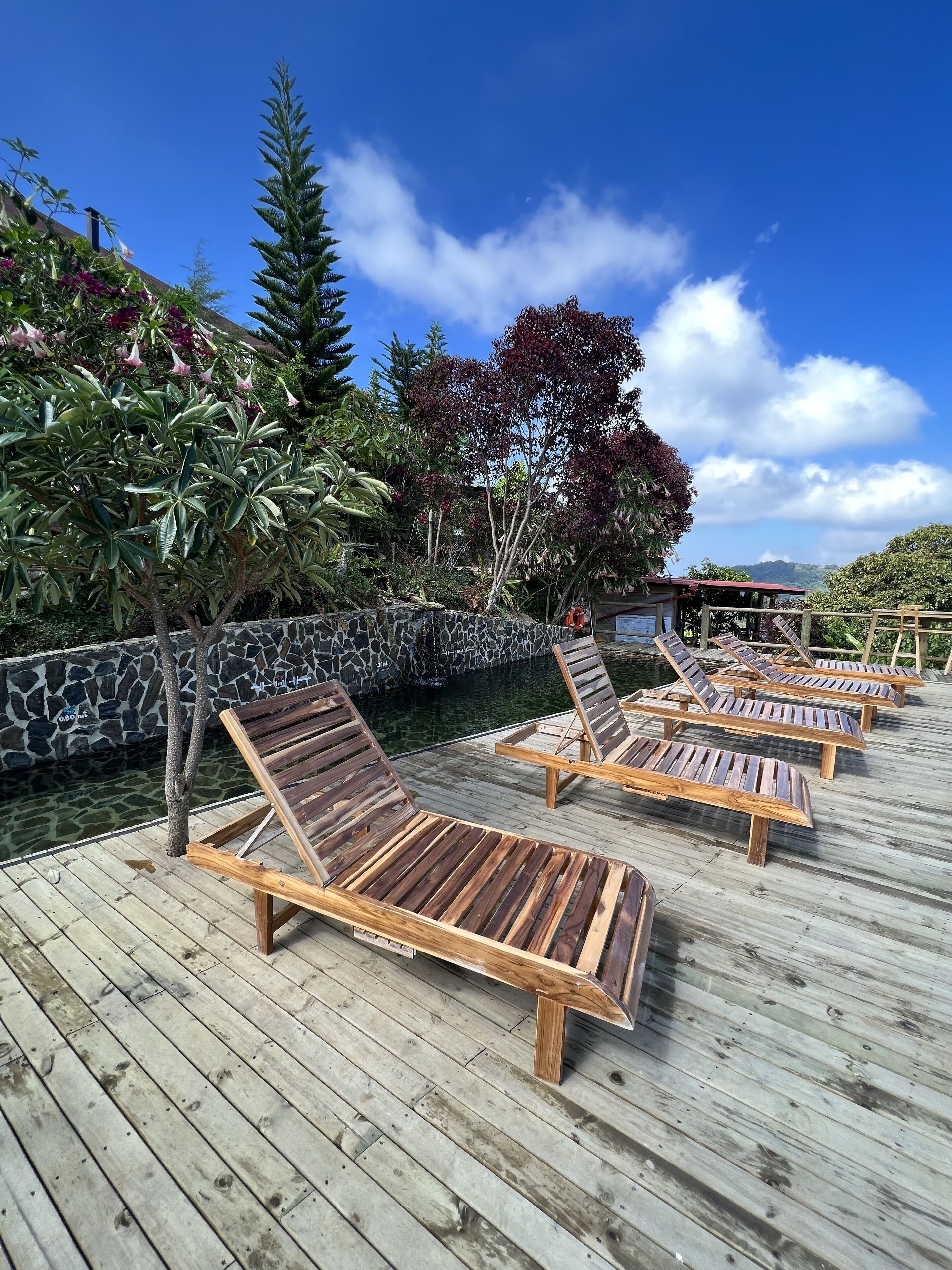 A wooden deck with lounge chairs, a stone wall, and a clear pool.