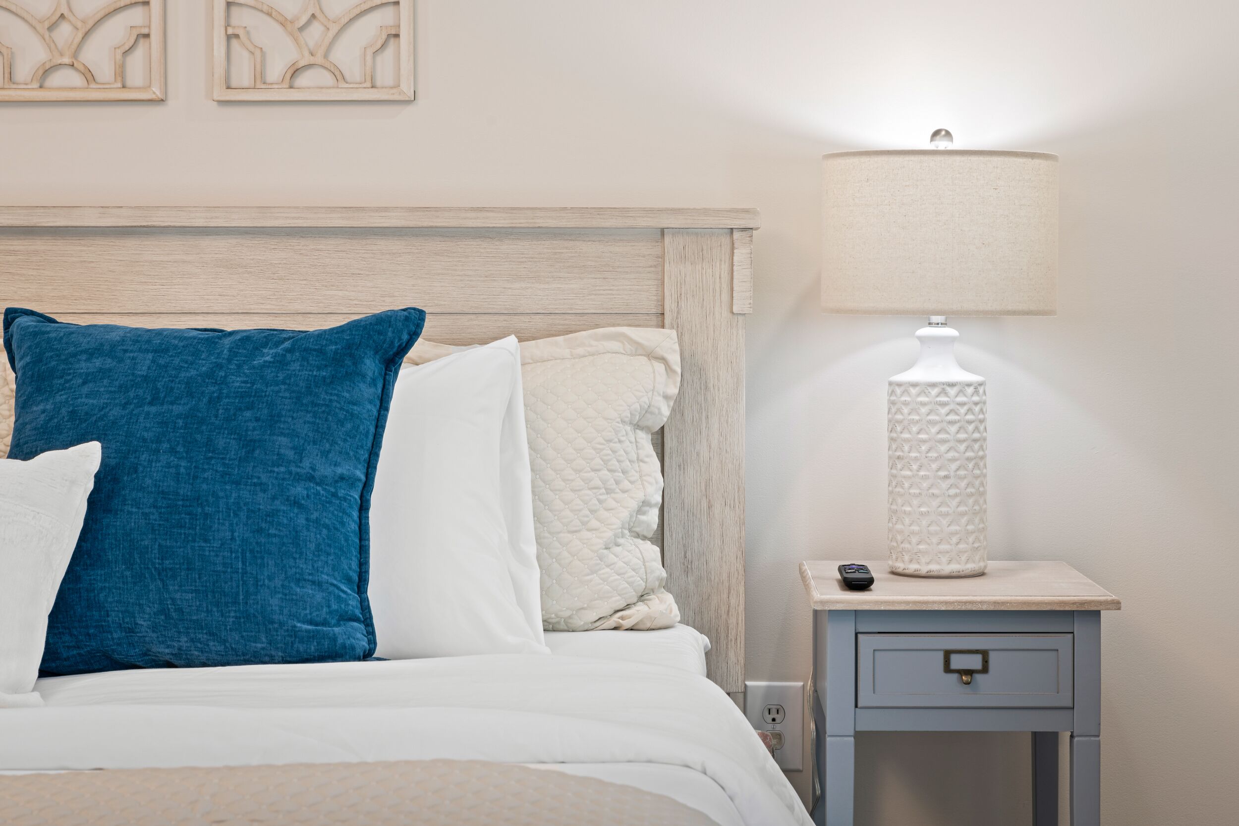 Un lit bien fait, avec un oreiller bleu et blanc, une couverture blanche matelassée et une lampe blanche posée sur une table de chevet.
