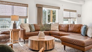 Un salon spacieux avec un canapé d’angle en cuir marron, une table basse ronde en bois et une table d’appoint avec une lampe. De grandes fenêtres laissent entrer la lumière naturelle et offrent une vue sur l’extérieur.
