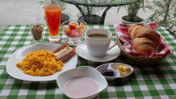 Desayuno con huevos revueltos, jamón, tostadas, fruta, café y jugo sobre un mantel a cuadros.
