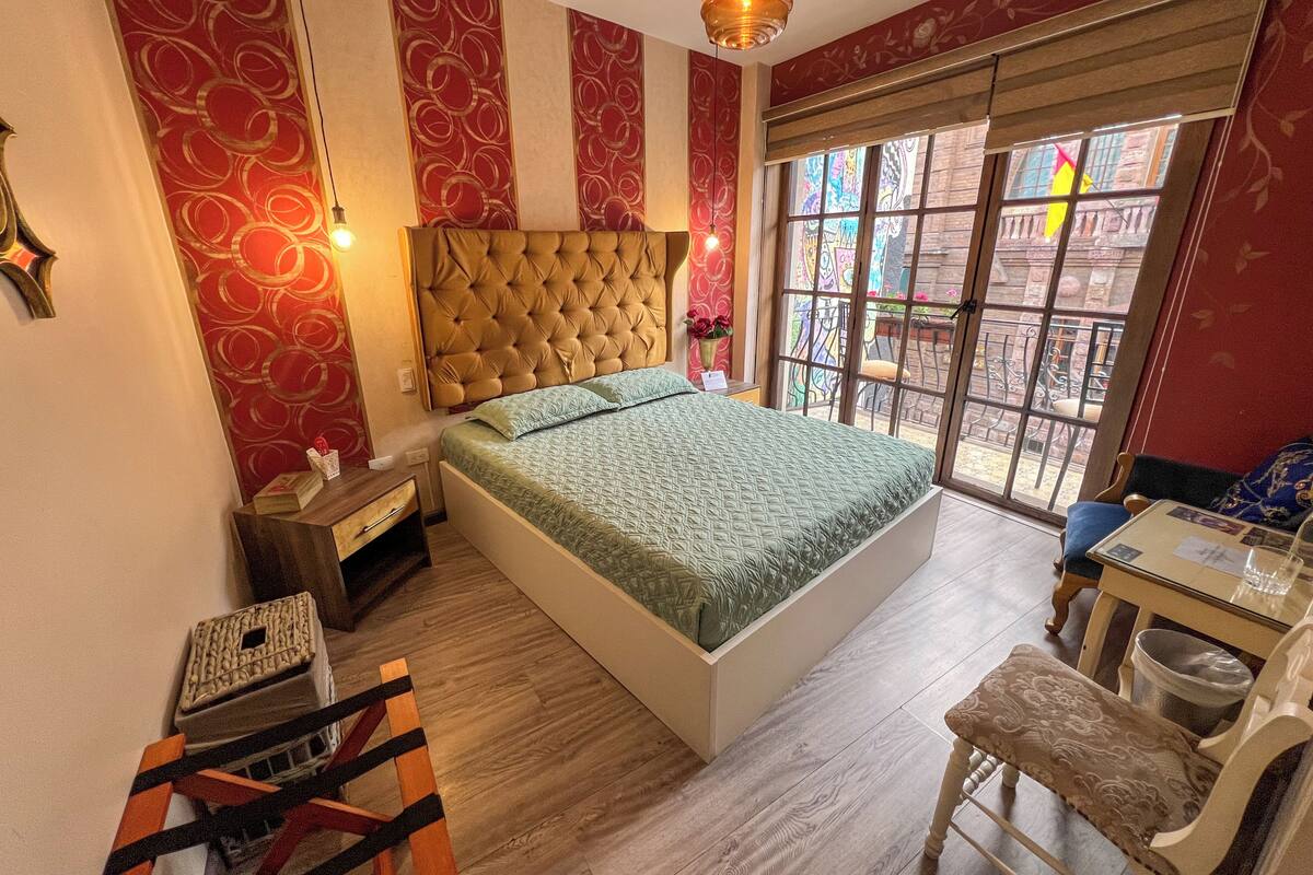 A bedroom with a bed, a desk, a chair, a window with a view, and a hanging light fixture.