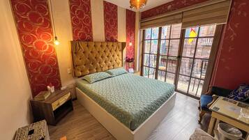 Un dormitorio con cabecera capitonada, una cama con ropa de cama estampada, una mesita de noche y una silla junto a una ventana con vistas, y una pequeña mesa.