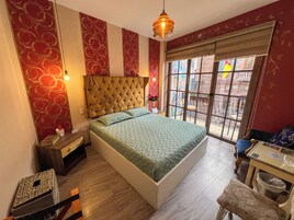 Un dormitorio con cabecera capitonada, una cama con ropa de cama estampada, una mesita de noche y una silla junto a una ventana con vistas, y una pequeña mesa.