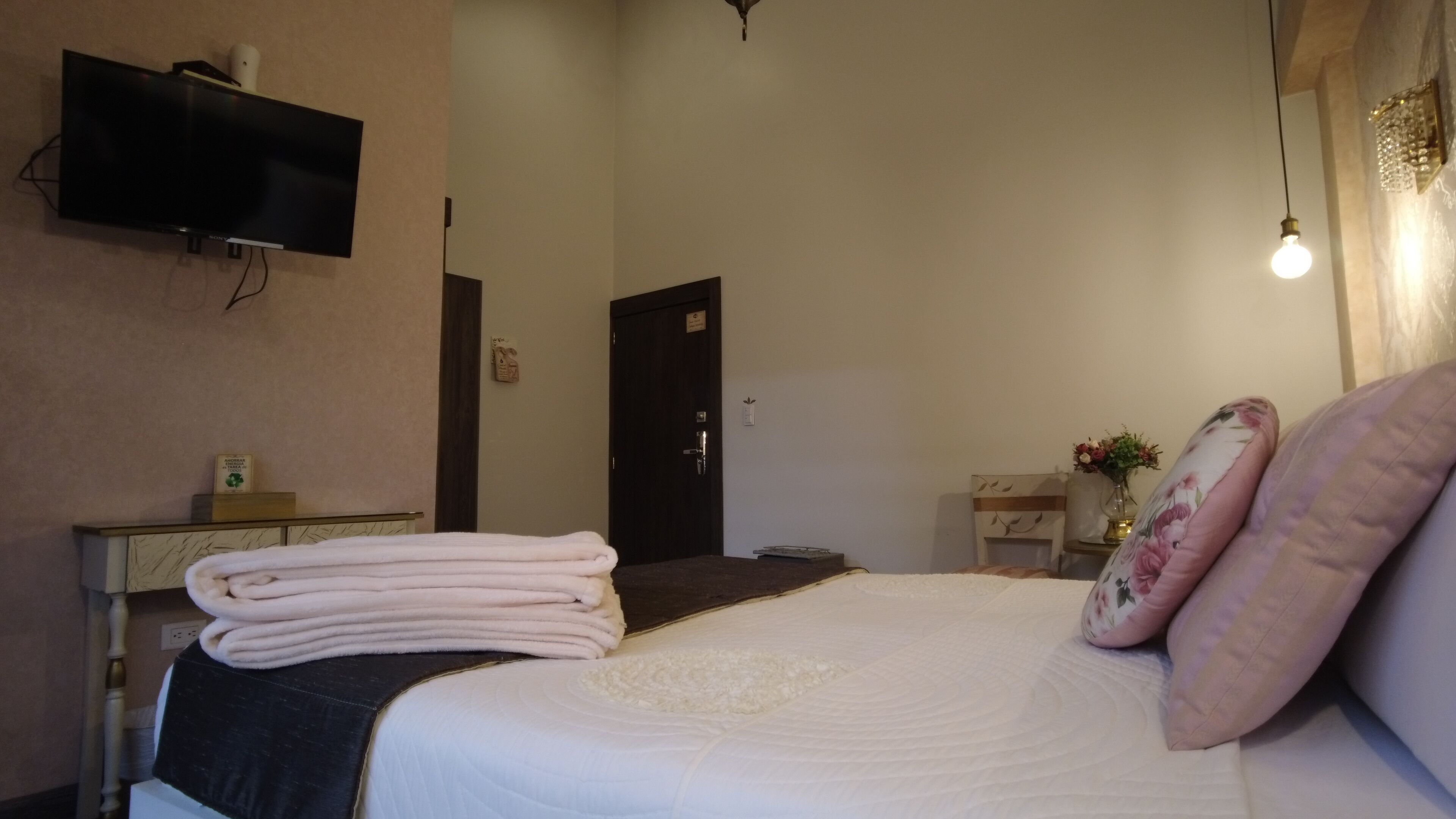 A hotel room with a bed, a flat-screen TV mounted on the wall, a desk with a lamp, and a door.