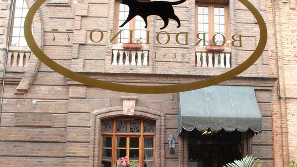 A breakfast setup with a checkered tablecloth, a glass of orange juice, a bowl of cereal, a plate of eggs, a cup, a basket of bread, and a potted plant in front of a building with a cat logo and the name "BORDONI" above the entrance. - Hotel Boutique La Ronda by Chat Noir (Cuenca)