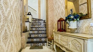 A hallway with patterned wallpaper, a tiled floor, a staircase with a decorative railing, a mirror with a gold frame, and a cabinet with a vase of flowers. - Hotel Boutique La Ronda by Chat Noir (Cuenca)
