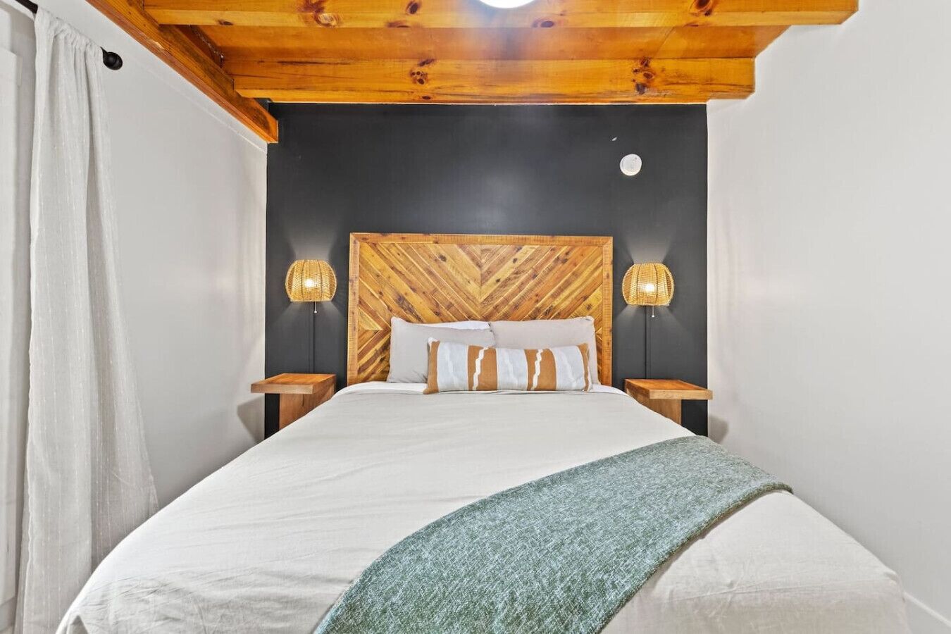 Un dormitorio con cabecera de madera, dos lámparas de noche, una cama con ropa blanca y un plaid verde, y una cortina blanca a la izquierda.