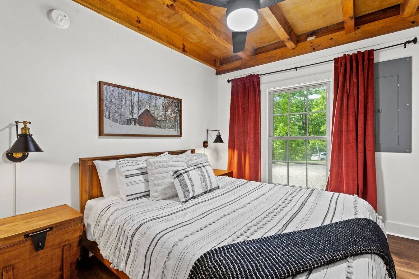 Une chambre avec un plafond en bois, un grand lit avec une literie à rayures, une fenêtre avec des rideaux rouges et un tableau accroché au mur.