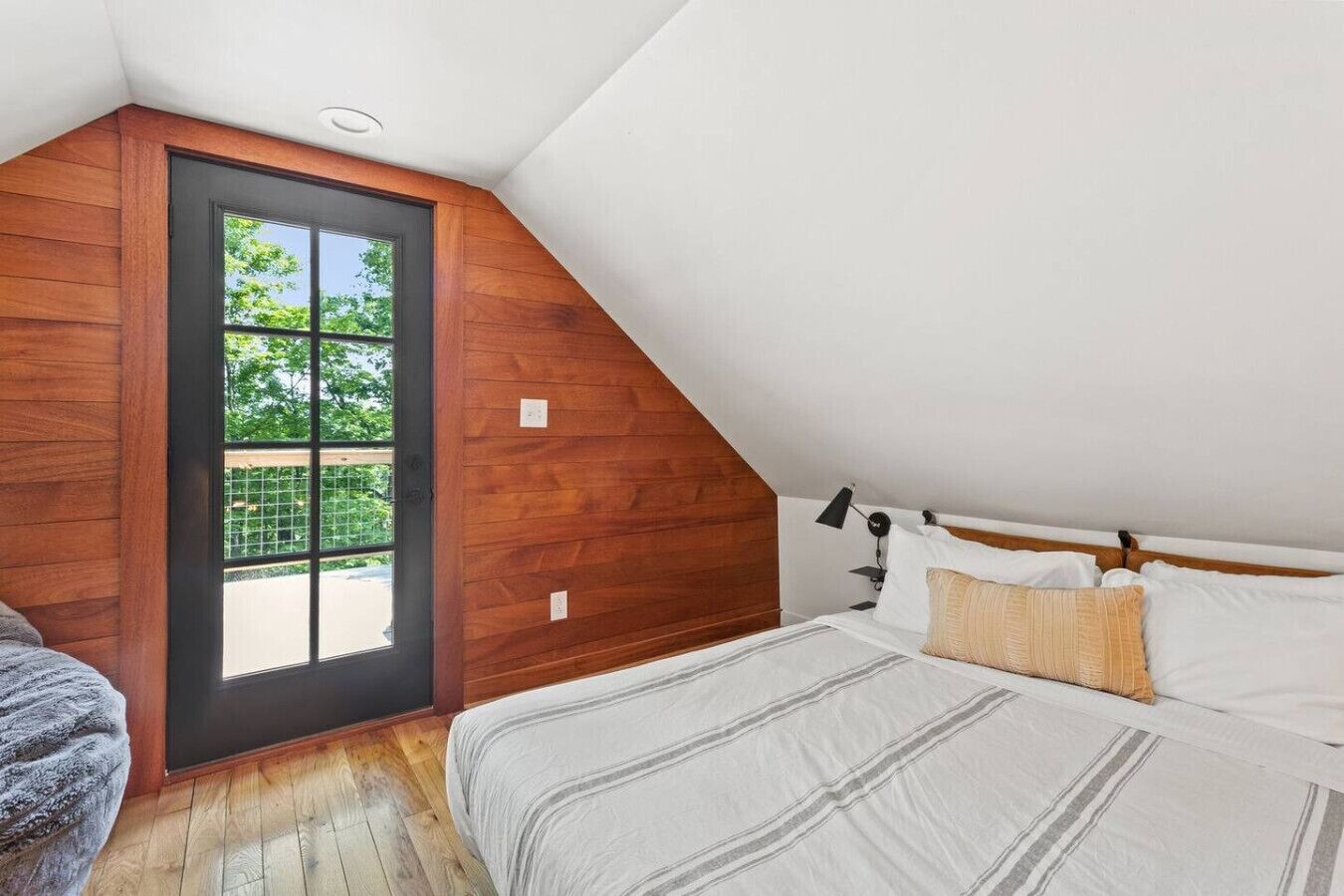 Een slaapkamer met een schuine kap, een groot raam, een bed met witte beddengoed en een houten vloer.