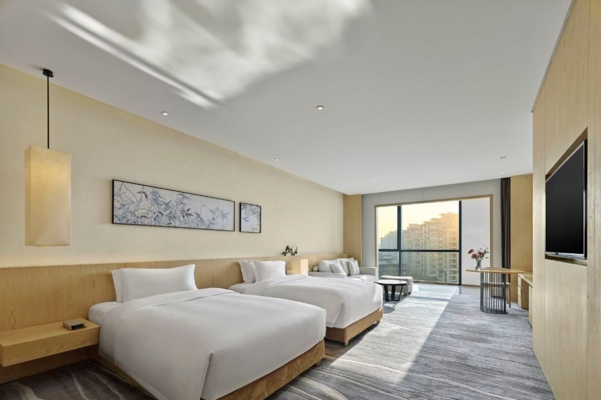 Habitación de hotel con dos camas, televisión, escritorio y un amplio ventanal con vistas a la ciudad.