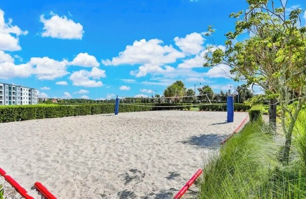 Una cancha de voleibol de playa con arena, una red, rodeada de follaje y un edificio al fondo.
