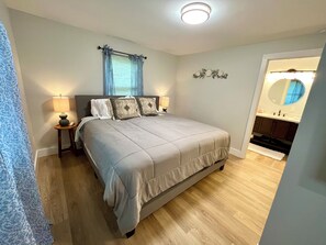 A bedroom with a bed, two bedside lamps, a window with blue curtains, a wall-mounted mirror, and a wooden floor.