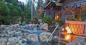 A hot spring area with large rocks, a wooden deck, and a building with a warm light.