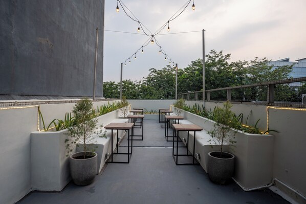 A rooftop terrace with tables and planters, string lights, and a view of trees and buildings.