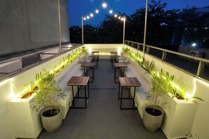 Un área de estar al aire libre con mesas y sillas, rodeada de macetas con plantas y luces colgantes.