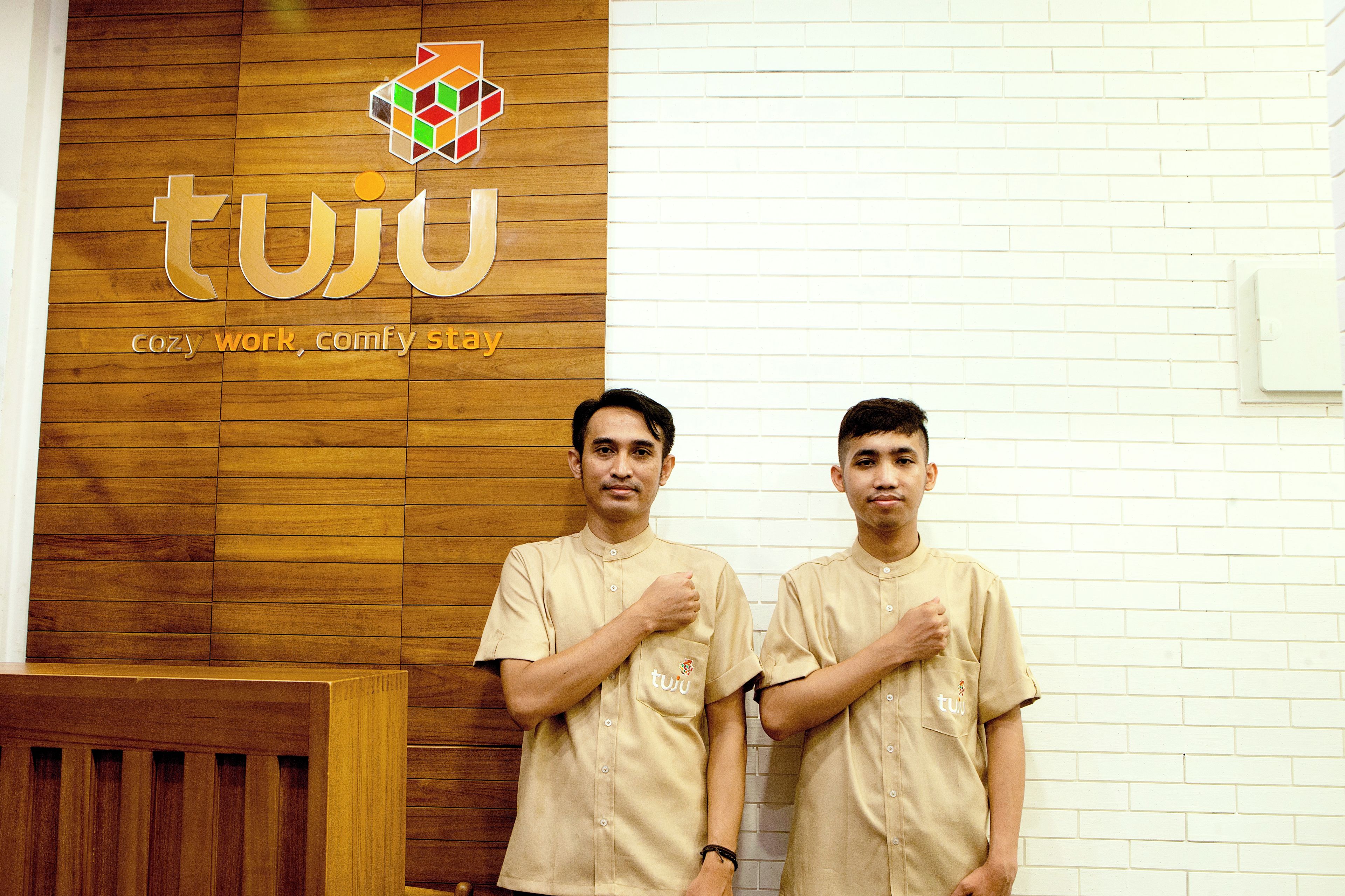 Two individuals standing in front of a wooden wall with a logo and text that reads 'tuju' and 'cozy work, comfy stay'.