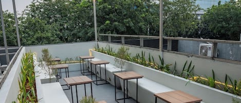 Een terras met tafels en banken, omringd door plantenbakken en lichtslingers.