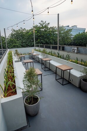 An outdoor seating area with tables and benches, surrounded by planters and string lights.