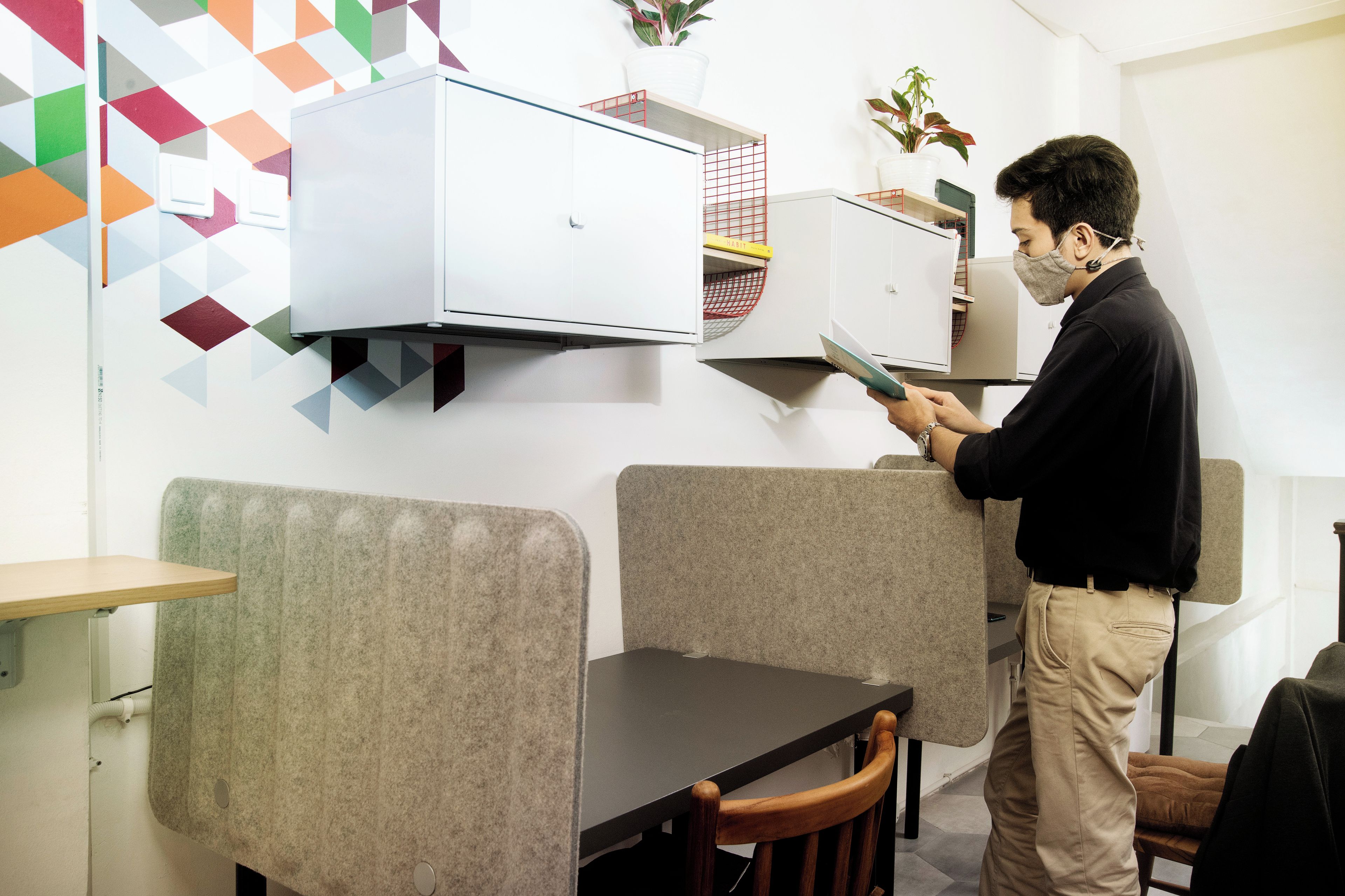 Une personne se tient dans une pièce qui contient une table et des chaises, un appareil de climatisation blanc et un mur orné de motifs géométriques.