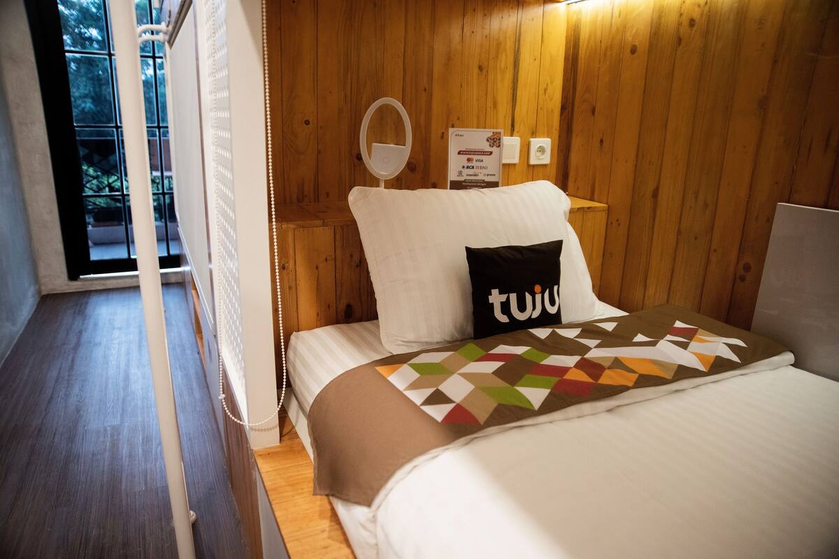 A hotel room with a wooden interior, a bed with a patterned blanket, a pillow with the word "tuju", a bedside table with a mirror, and a sign indicating payment options.