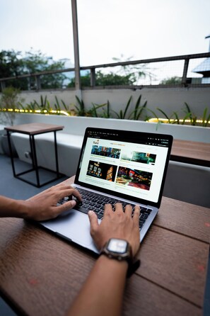 A person using a laptop on a wooden table with a view of greenery and a building in the background. - Tuju Arteri Pods by Zuzu (Jakarta)