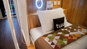 A hotel room with a wooden wall, a bed with a geometric-patterned blanket, a white pillow, a black pillow with the word "tuju," a bedside table with a ring light, and a sign indicating payment options. - Tuju Arteri Pods by Zuzu (Jakarta)