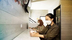 Two individuals seated at a marble table in a modern interior with a wooden wall and a television mounted above. - Tuju Arteri Pods by Zuzu (Jakarta)