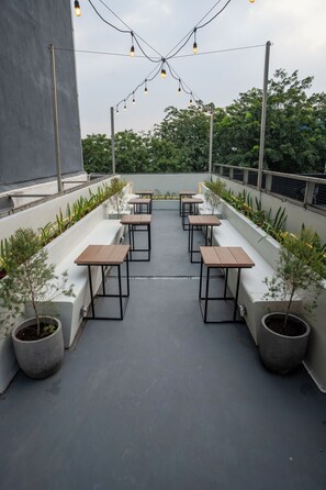 Un área exterior con mesas y sillas, rodeada de plantas en macetas y luces colgantes.