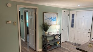 A hallway with a white door, a wall-mounted thermostat, a small potted plant, a framed map, and a table with a lamp and a clock.