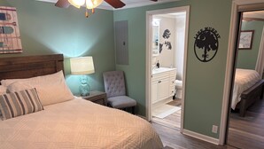 A bedroom with a bed, nightstand, lamp, and a chair. There is a bathroom visible through an open door.