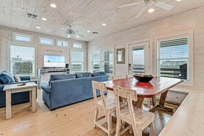 Un salón espacioso con techo de madera, un ventanal grande, un sofá azul, una mesa de comedor con sillas y un televisor.