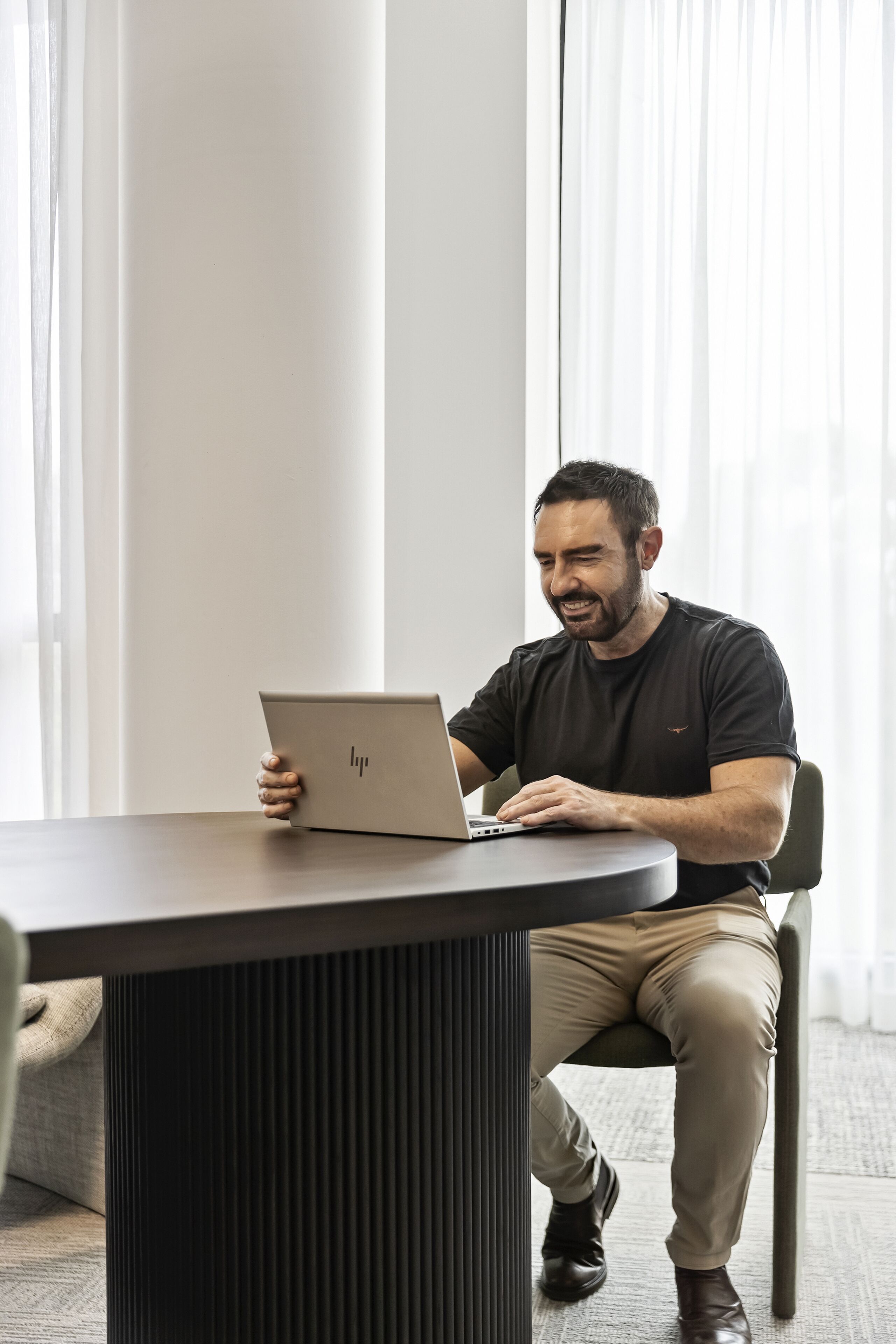 A person seated at a table with a laptop, in a room with large windows and a modern design.